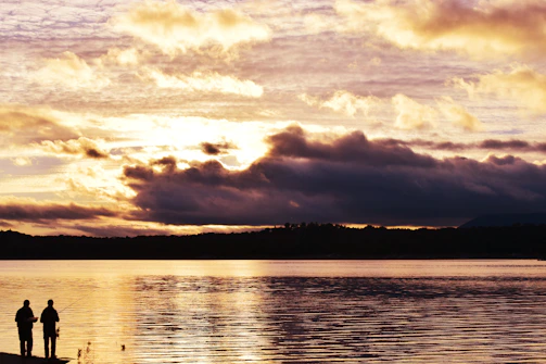 A rugged outdoor scene with a hunter and angler exchanging a friendly handshake near a forest lake at sunset.