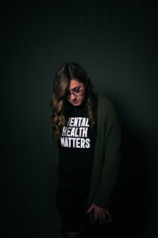 A person wearing glasses and a shirt with the phrase 'Mental Health Matters' printed on it stands against a dark background. They have long, wavy hair and are wearing a green cardigan.