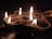 A cluster of small clay lamps, traditionally known as diyas, are lit and arranged on a dark surface. The warm glow of the flames creates a soft, serene atmosphere. The background is dimly lit, highlighting the gentle flicker of the fire against the clay texture.