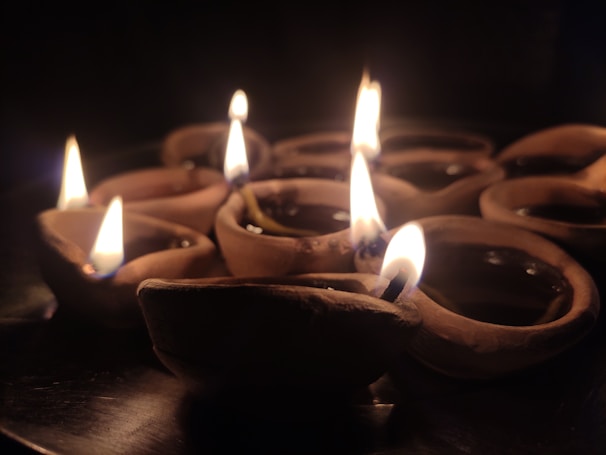 A cluster of small clay lamps, traditionally known as diyas, are lit and arranged on a dark surface. The warm glow of the flames creates a soft, serene atmosphere. The background is dimly lit, highlighting the gentle flicker of the fire against the clay texture.
