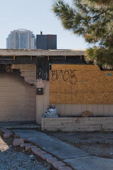 A partially burned, abandoned building has boarded-up windows with graffiti on them. Next to the charred area is a small pile of debris. A tall modern building is visible in the background, and a tree with green foliage is on the right side of the image.