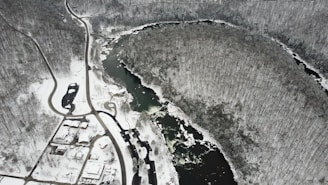 An aerial view of a snow-covered landscape featuring a winding river surrounded by dense, leafless trees. Roads and a few buildings are visible in the lower portion of the image, with a stark contrast between the dark water and the white snow.