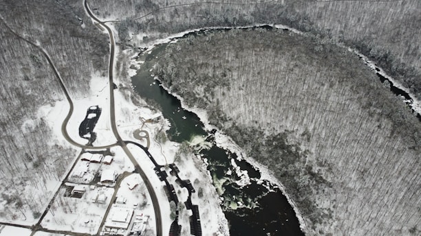 An aerial view of a snow-covered landscape featuring a winding river surrounded by dense, leafless trees. Roads and a few buildings are visible in the lower portion of the image, with a stark contrast between the dark water and the white snow.