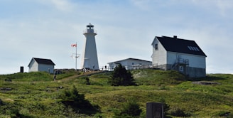 A picturesque scene depicting a white lighthouse with a red Canadian flag waving nearby, set on a grassy hill. The surrounding area includes several white buildings with dark roofs. The sky is clear and blue.