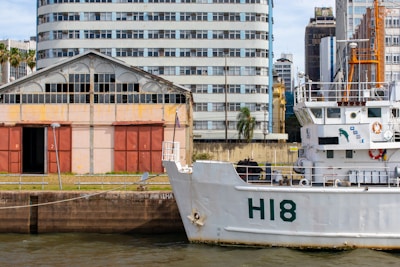 A maritime scene featuring a large white ship docked near an industrial area. The ship has 'H18' marked on its hull and various equipment and life rings visible on its decks. Behind the ship, there is an old warehouse with red doors and a weathered facade, alongside an array of urban buildings of varying architectural styles. Palm trees and patches of greenery are integrated into the urban landscape.