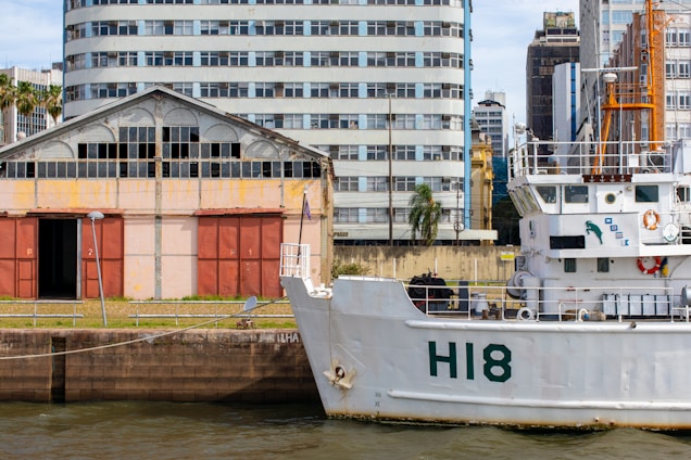 A maritime scene featuring a large white ship docked near an industrial area. The ship has 'H18' marked on its hull and various equipment and life rings visible on its decks. Behind the ship, there is an old warehouse with red doors and a weathered facade, alongside an array of urban buildings of varying architectural styles. Palm trees and patches of greenery are integrated into the urban landscape.