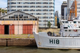 A maritime scene featuring a large white ship docked near an industrial area. The ship has 'H18' marked on its hull and various equipment and life rings visible on its decks. Behind the ship, there is an old warehouse with red doors and a weathered facade, alongside an array of urban buildings of varying architectural styles. Palm trees and patches of greenery are integrated into the urban landscape.