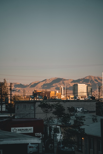 An urban landscape with a foreground of buildings and a large truck labeled 'Republic Services'. In the background, mountains are visible under a clear blue sky, with the sun casting a warm glow on the structures.