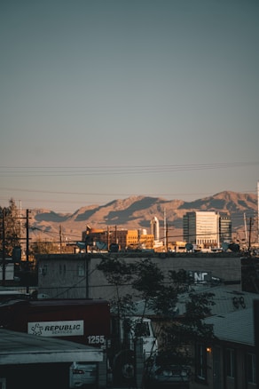 An urban landscape with a foreground of buildings and a large truck labeled 'Republic Services'. In the background, mountains are visible under a clear blue sky, with the sun casting a warm glow on the structures.