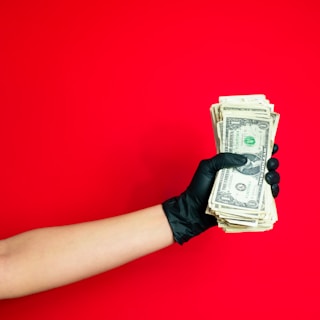 An arm wearing a black glove holds a stack of U.S. dollar bills against a bright red background.