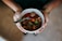 A pair of hands holding a white bowl filled with a hearty stew containing large chunks of meat, cherry tomatoes, and sliced green peppers. A metal spoon is placed inside the bowl, ready for use. The person wearing a gold wristwatch stands against a blurred background.