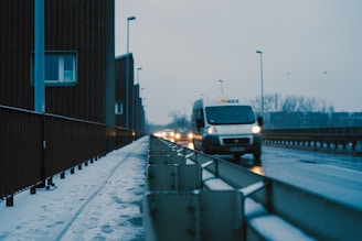 A snowy street in a city with a parked van on the right side. The van has a company logo on it, and there are faint tire tracks in the snow. A building with a window is on the left, while streetlights line the road. The sky is overcast, and the atmosphere appears cold and quiet.