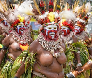 A group of people dressed in traditional attire with elaborate headdresses made of feathers, shells, and vibrant face paint. They are adorned with necklaces made of shells and leaves, and their expressions convey excitement and joy. The attire includes grass skirts and decorative arm bands.