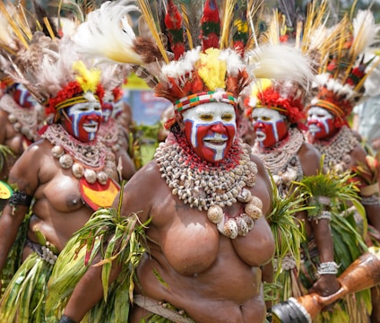 A group of people dressed in traditional attire with elaborate headdresses made of feathers, shells, and vibrant face paint. They are adorned with necklaces made of shells and leaves, and their expressions convey excitement and joy. The attire includes grass skirts and decorative arm bands.