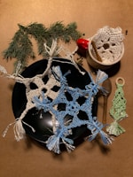 Crocheted snowflake motifs with light blue and white threads are arranged on a dark plate, accompanied by a small crocheted green tree. Next to the plate, a small white ceramic bowl contains a red bell and a cinnamon stick. A sprig of evergreen branches adds a natural touch to the scene.