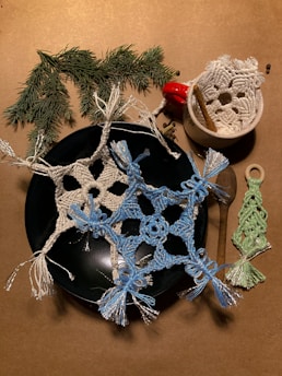 Crocheted snowflake motifs with light blue and white threads are arranged on a dark plate, accompanied by a small crocheted green tree. Next to the plate, a small white ceramic bowl contains a red bell and a cinnamon stick. A sprig of evergreen branches adds a natural touch to the scene.