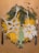 Intricate crochet ornaments in white and light green are arranged on a vibrant yellow textured mat. Pine branches are positioned around, creating a natural frame. Accompanying the composition is a set of silverware and scattered peppercorns with a cinnamon stick, all placed on a brown background.