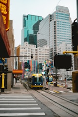 A modern cityscape featuring tall glass and steel skyscrapers with a light rail train moving along the tracks in the foreground. The scene includes colorful street art murals on nearby buildings and a pedestrian crossing. The architecture and urban setting create a bustling, lively atmosphere.