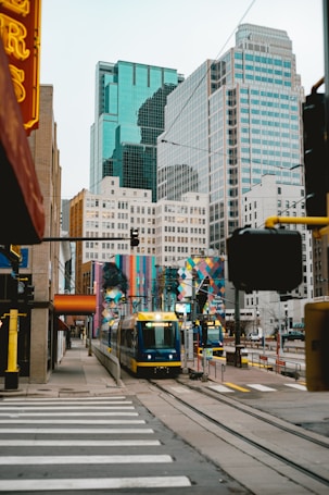 A modern cityscape featuring tall glass and steel skyscrapers with a light rail train moving along the tracks in the foreground. The scene includes colorful street art murals on nearby buildings and a pedestrian crossing. The architecture and urban setting create a bustling, lively atmosphere.