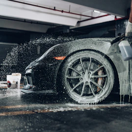 A person spraying soapy foam onto a black sports car in a garage
