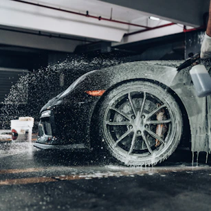 A person spraying soapy foam onto a black sports car in a garage