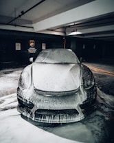 A sleek car covered in foamy soap is parked in a dimly lit garage, with signs indicating no parking and steam cleaning services on the wall in the background.