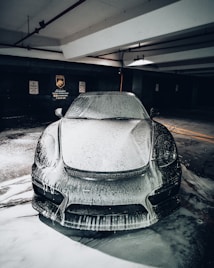 A sleek car covered in foamy soap is parked in a dimly lit garage, with signs indicating no parking and steam cleaning services on the wall in the background.