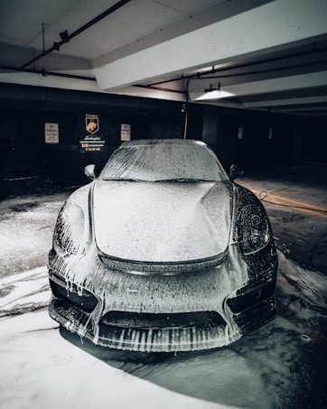 A sleek car covered in foamy soap is parked in a dimly lit garage, with signs indicating no parking and steam cleaning services on the wall in the background.