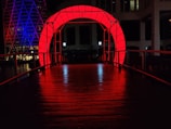 A bridge is illuminated by vibrant red lighting, creating a striking archway. In the background, a building with geometric patterns is lit in purple and blue hues. The scene is at night, enhancing the contrast between the bright lights and the darkness.