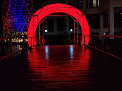 A bridge is illuminated by vibrant red lighting, creating a striking archway. In the background, a building with geometric patterns is lit in purple and blue hues. The scene is at night, enhancing the contrast between the bright lights and the darkness.
