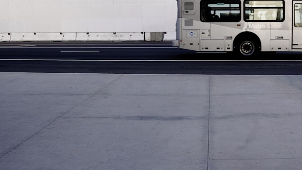 A mostly empty urban street scene with part of a public bus on the right side. The street is wide and bordered by a concrete barrier. The bus, marked with a few logos and the words 'Metro Local', is beige with black windows and wheels.
