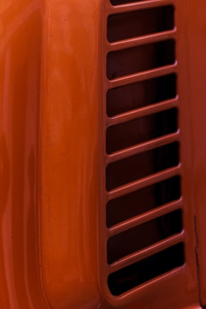 A close-up view of a bright orange surface with parallel rectangular ventilation slits. The surface has a glossy finish with some visible imperfections or speckles.