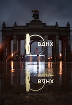 An illuminated sign with Cyrillic letters is prominently displayed in front of a grand archway with statues on top. The scene is reflected on a wet surface, creating a mirror effect. The lighting is moody, with a dark, cloudy sky overhead.