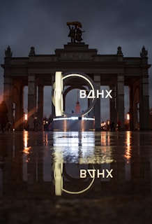 An illuminated sign with Cyrillic letters is prominently displayed in front of a grand archway with statues on top. The scene is reflected on a wet surface, creating a mirror effect. The lighting is moody, with a dark, cloudy sky overhead.