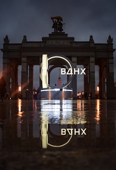 An illuminated sign with Cyrillic letters is prominently displayed in front of a grand archway with statues on top. The scene is reflected on a wet surface, creating a mirror effect. The lighting is moody, with a dark, cloudy sky overhead.