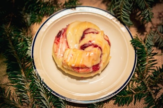 A glazed pastry with a swirled shape sits on a white plate with a dark rim. The pastry has a glossy finish, indicating freshness, with sections of red filling visible, likely fruit or jam. Surrounding the plate, there are green pine branches, suggesting a festive or holiday setting.