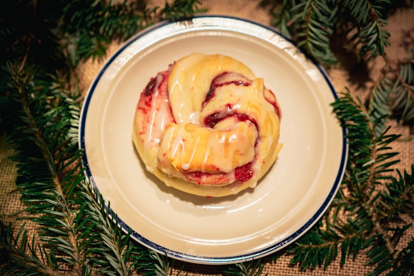 A glazed pastry with a swirled shape sits on a white plate with a dark rim. The pastry has a glossy finish, indicating freshness, with sections of red filling visible, likely fruit or jam. Surrounding the plate, there are green pine branches, suggesting a festive or holiday setting.