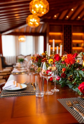 An elegantly set dining table features a lush floral arrangement with a variety of flowers in reds and oranges. The table is set with neatly arranged cutlery, wine glasses, and place settings atop woven placemats. The warm glow from the overhead geometric pendant lights creates a cozy and inviting atmosphere. The room's decor is sophisticated, showcasing wooden beams and a softly lit background.