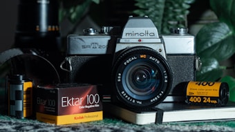 A vintage Minolta SR-T 200 camera is positioned at the center, surrounded by film rolls and boxes of Kodak Professional 35mm color negative film, including Ektar 100 and Kodak 400. The scene includes a notebook and a partially visible camera film roll, set against a backdrop of green leaves, suggesting a nostalgic, analog photography theme.