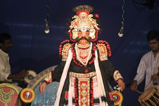 A performer is dressed in elaborate traditional attire with intricate designs and vibrant colors, including a detailed headdress, face paint, and a richly decorated costume. Two musicians are also present, one playing drums, and the other appears to be preparing an instrument. The background is a deep blue, and the scene appears to be part of a cultural or theatrical performance.