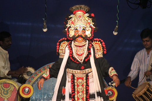 A performer is dressed in elaborate traditional attire with intricate designs and vibrant colors, including a detailed headdress, face paint, and a richly decorated costume. Two musicians are also present, one playing drums, and the other appears to be preparing an instrument. The background is a deep blue, and the scene appears to be part of a cultural or theatrical performance.