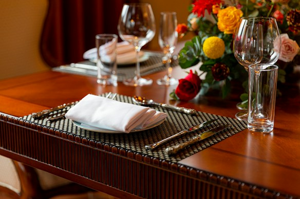 A warmly lit dining table is elegantly set with a woven placemat, silver cutlery, a folded white napkin, and clear glassware. A vibrant floral arrangement with red, yellow, and pink flowers adds a touch of color to the scene.