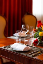 A dining table set elegantly with folded white napkins on white plates, surrounded by cutlery and wine glasses. A vibrant floral arrangement with red, yellow, and orange flowers is partially visible, enhancing the decor. The background features plush chairs and red curtains, giving the setting a warm and luxurious ambiance.