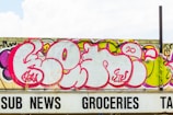 A large graffiti mural with bold pink, purple, and white lettering covers a concrete wall. The artwork features stylized, rounded letters with intricate details in a contemporary urban art style. Below the wall is a partially visible sign with black lettering spelling 'SUB NEWS GROCERIES TAX.'