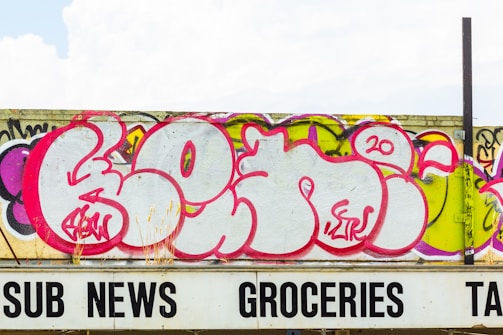 A large graffiti mural with bold pink, purple, and white lettering covers a concrete wall. The artwork features stylized, rounded letters with intricate details in a contemporary urban art style. Below the wall is a partially visible sign with black lettering spelling 'SUB NEWS GROCERIES TAX.'