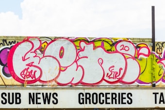A large graffiti mural with bold pink, purple, and white lettering covers a concrete wall. The artwork features stylized, rounded letters with intricate details in a contemporary urban art style. Below the wall is a partially visible sign with black lettering spelling 'SUB NEWS GROCERIES TAX.'