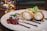 A dessert is elegantly plated with two crispy pastry rolls filled with a creamy filling, drizzled with chocolate sauce. Beside them lie red berries and a sprig of mint. The background includes a softly lit candle and blurred books.
