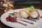 A dessert is elegantly plated with two crispy pastry rolls filled with a creamy filling, drizzled with chocolate sauce. Beside them lie red berries and a sprig of mint. The background includes a softly lit candle and blurred books.
