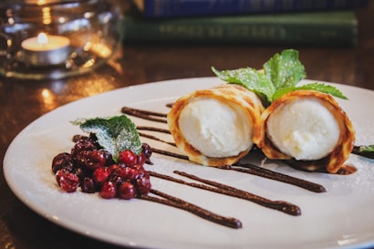 A dessert is elegantly plated with two crispy pastry rolls filled with a creamy filling, drizzled with chocolate sauce. Beside them lie red berries and a sprig of mint. The background includes a softly lit candle and blurred books.