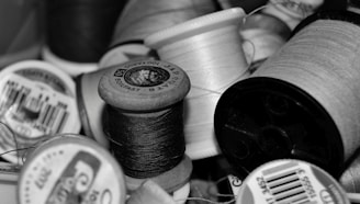 A collection of thread spools in various sizes and colors, close-up. The threads are tightly wound around the spools, and some have labels with brand names and product details. The arrangement appears random, showing a variety of textures and materials.